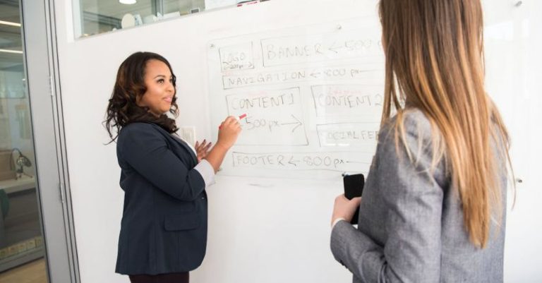 Teaching - Woman in Black Blazer Looking at Woman in Grey Blazer