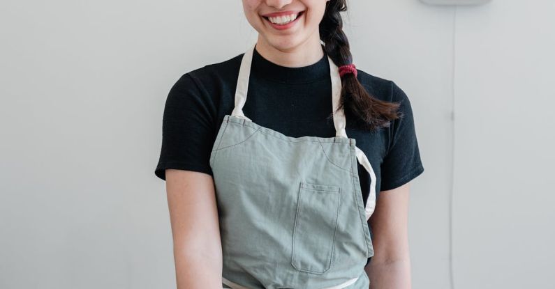 Job Offer - Positive young Asian woman wearing apron giving paper cup with coffee to go while standing at wooden counter during work in modern coffee house and looking at camera with smile
